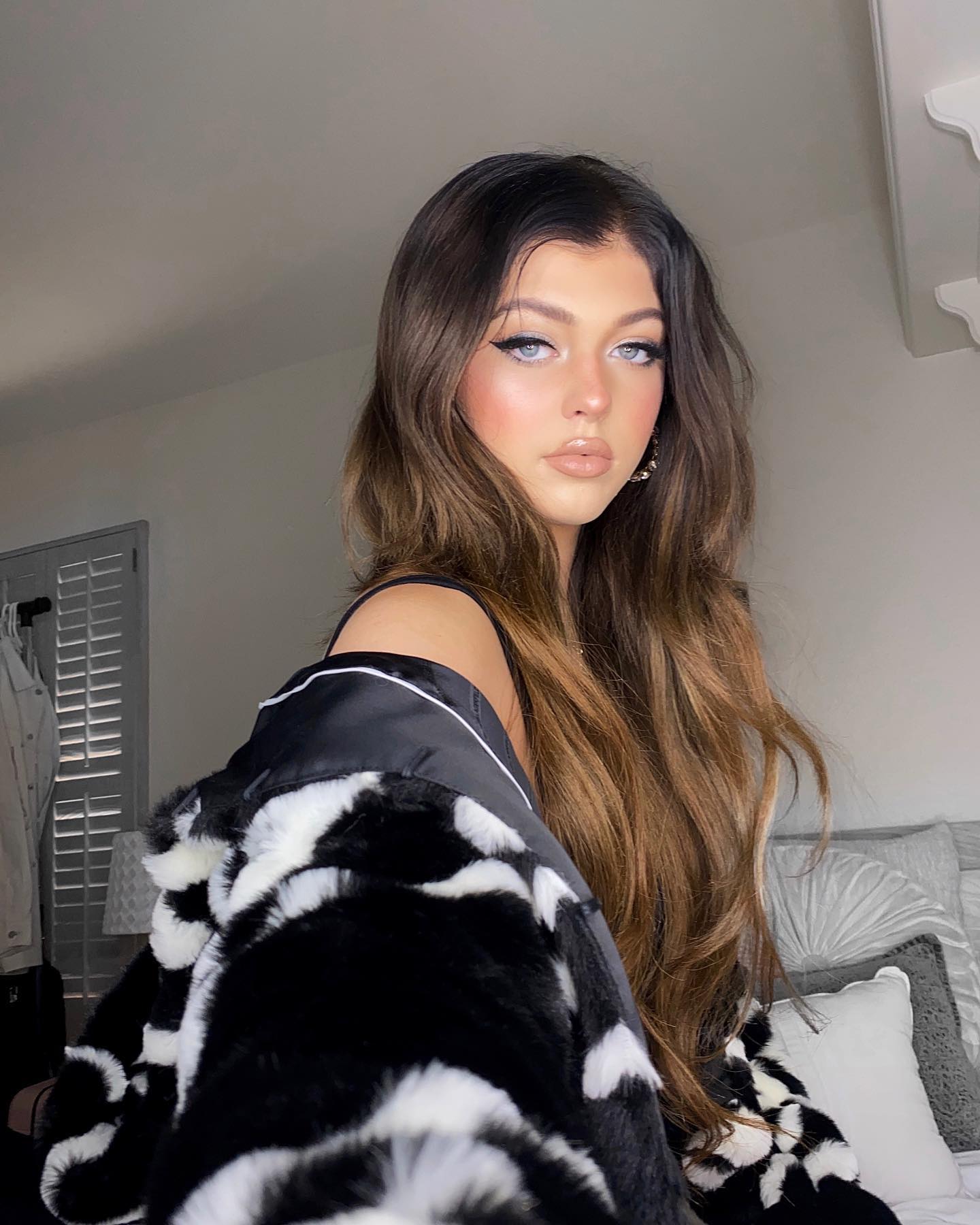 a woman with long, wavy brunette hair and fair skin is posing for a picture. she is wearing a black tank top and a black and white fur coat. her makeup includes white eyeliner, blush, and nude lipstick. she is standing in a bedroom with a bed and pillows in the background. shutters can also be seen in the background.
