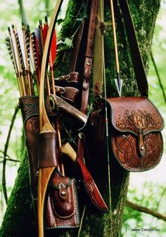 the image shows a recurve bow and arrow set leaning against a tree in a forest. the bow is made of wood and has a leather grip. the arrows are in a leather quiver that is decorated with tooling. the quiver is attached to a leather belt that also has a pouch. the background of the image is a blurry green forest.