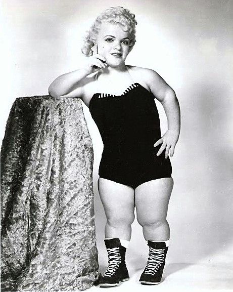 the black and white photograph shows a female little person, dressed in a black swimsuit and highlaced boots, posing confidently.  she is leaning against a textured, draped fabric backdrop. her hair is styled in a bouffant. the setting appears to be a professional studio. the image likely depicts a wrestler or performer from a mid20th century circus or sideshow.  the overall tone is one of strength and selfassuredness despite the potentially challenging circumstances.