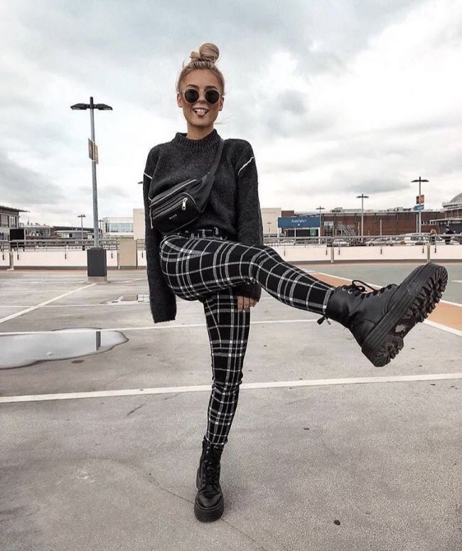 a full shot shows a woman standing on a paved surface, possibly a parking lot or large outdoor area, with buildings and light posts visible in the background. she is dressed in a grey sweater, black and white plaid pants, and black boots. she is wearing black sunglasses and has her hair styled in a high bun. a black cross body bag is slung across her chest. the woman is striking a pose with one leg raised, and she is smiling, creating a playful and fashionable scene.