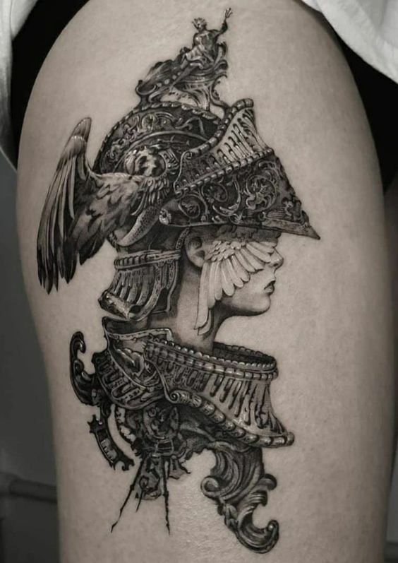 this black and grey tattoo depicts a woman in ornate armor. the detailed design showcases a helmet with large, feathered wings and intricate details. her expression is stoic and determined, hinting at an inner strength.  the overall aesthetic blends realism with a touch of fantasy.