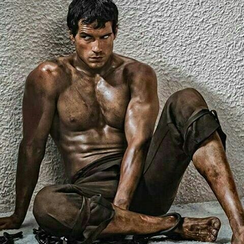 a shirtless man with a muscular physique sits with his legs crossed while his hands are bound with chains. he is sitting in front of a gray wall and appears to be posing for a photo shoot.