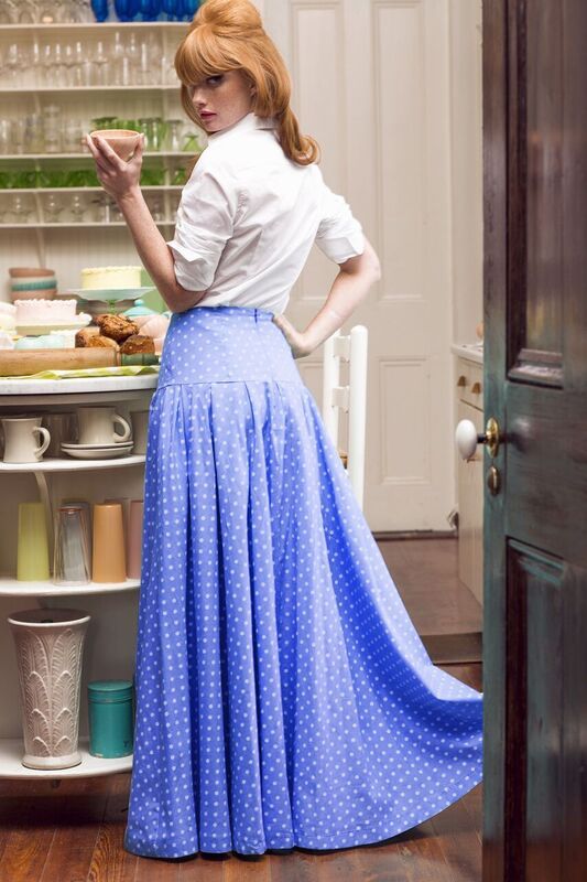 a woman in a vintageinspired outfit stands in a kitchen. she is wearing a white blouse and a long blue skirt with white polka dots. the skirt is highwaisted and flows to the floor. she has red hair styled in a vintage 'do. she is holding a teacup and looking over her shoulder. the kitchen is decorated in a retro style with pastel colors and vintage appliances. the lighting is soft and natural.