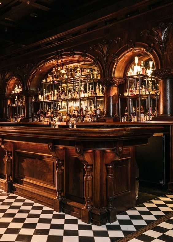 a shot of a dimly lit, luxurious bar with a dark wood counter and ornate detailing. behind the counter are shelves filled with liquor bottles and a checkered floor in black and white. the bar has a classic, vintage, speakeasy feel with a warm, intimate ambiance.