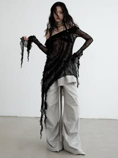a woman wearing a black sheer top with a white bottom is standing in a studio with her body angled to the left. the black top has lace detailing and drapes across the front of her body.  the top is long sleeved.  the woman has black hair, dark eyes, and pale skin.  she is wearing a necklace and has a bracelet on her left wrist. her pose is relaxed with arms slightly bent.