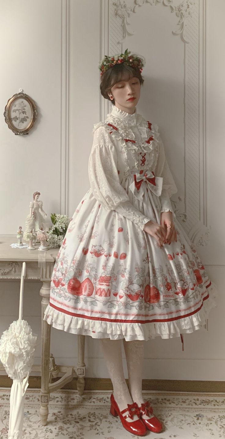 a young woman in a white and red lolita fashion dress stands in a room. she is wearing a white blouse with lace trim and a white and red strawberryprint skirt with a red bow. she has short brown hair with a flower crown and is wearing red shoes. the room is decorated in a vintage style with a white wall and a white table. there is a framed picture on the wall and a white umbrella leaning against the table. the lighting is soft and natural, and the overall effect is romantic and feminine.