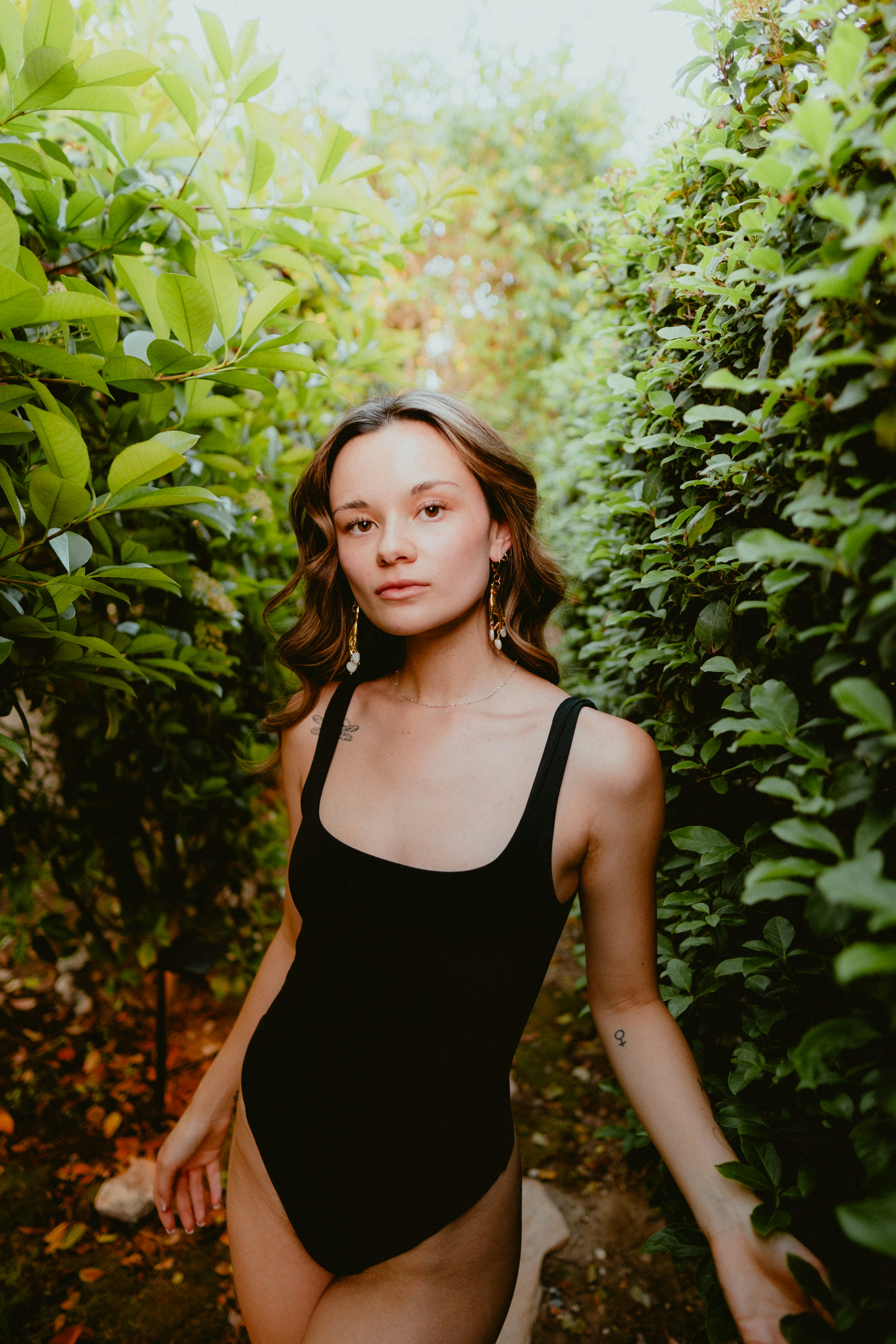 a young woman with long brown wavy hair stands in a path between two lush green hedges. she is wearing a black onepiece swimsuit and delicate earrings. she has a small tattoo on her forearm. the setting is outdoors, with sunlight filtering through the leaves. the woman is looking directly at the camera with a confident expression.  the image has a natural and slightly sensual feel.