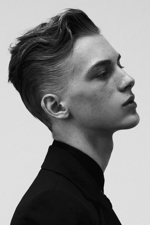 a black and white closeup profile portrait of a young man with a stylish, wavy, slickedback hairstyle. he's wearing a dark suit jacket with a high collar.  the image is sharply focused, highlighting his facial features and the texture of his hair. the overall mood is serious and elegant.