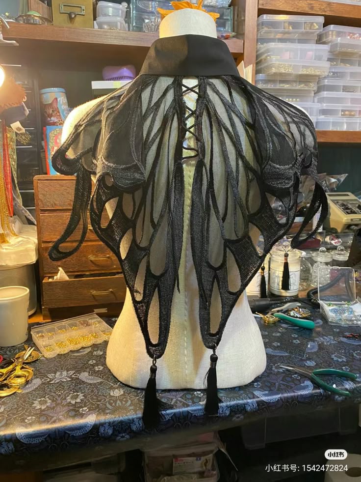 a black butterfly wing collar is displayed on a mannequin, featuring intricate designs and laceup detailing in the back, with tassels at the tips of the wings. it sits amidst a crafter's setup.