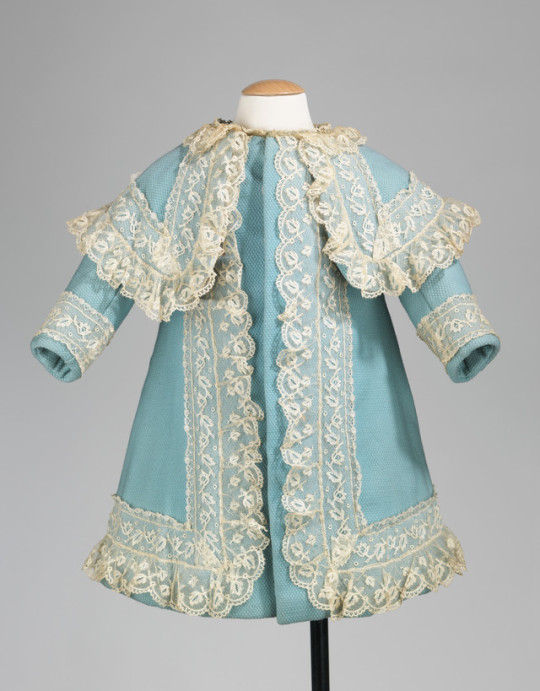 a front view of a baby blue baby dress with creamcolored lace accents. the dress is on a mannequin in front of a solid gray background.