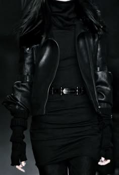 a young woman wearing a long black leather jacket, a black shirt, and a black belt poses for the camera. she has long black hair, and gloves.  the lighting is dark, creating a mysterious and edgy feel.