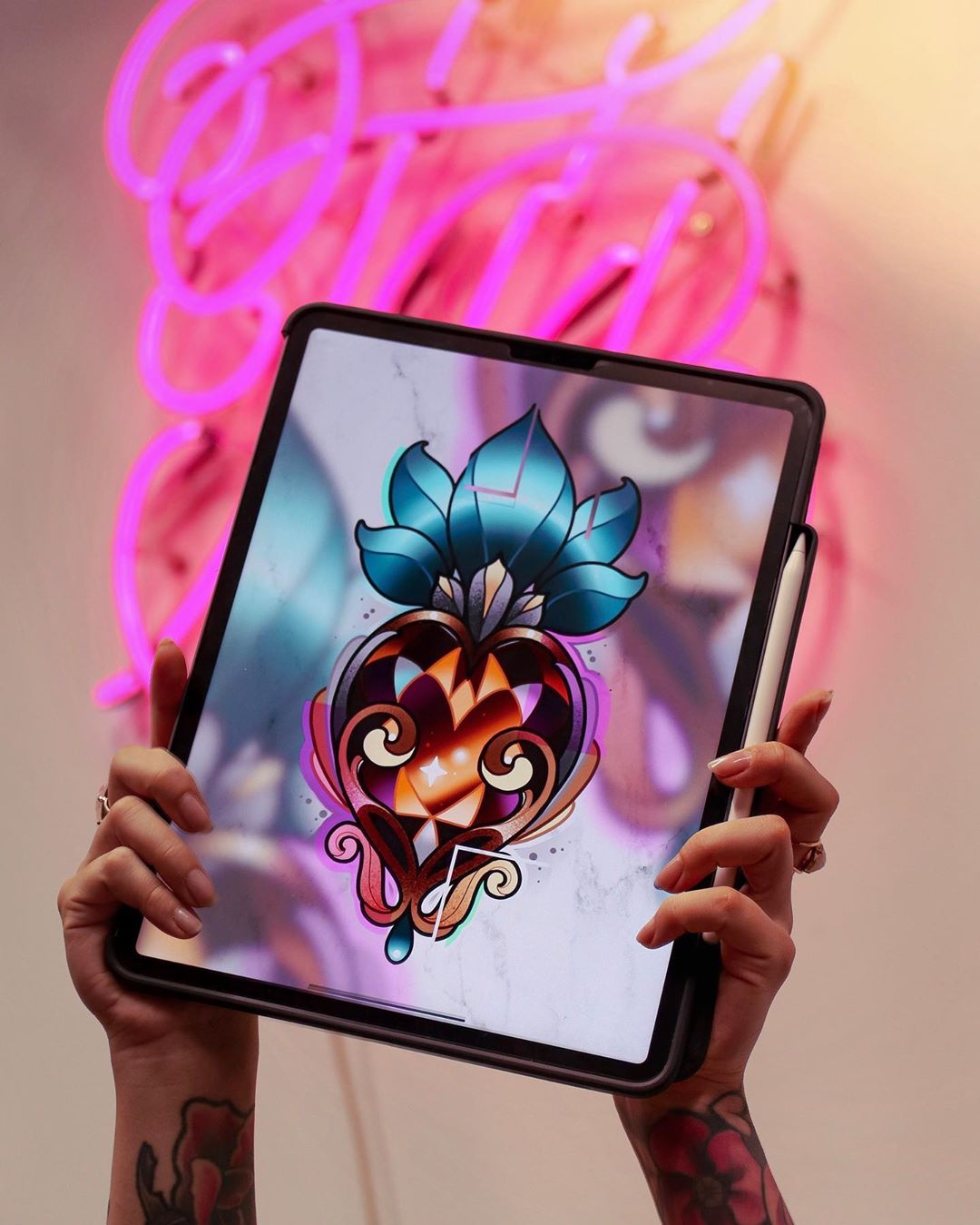 a woman holds up a tablet displaying a digital drawing of an ornately decorated pineapple with blue leaves against a pink neon sign in the background. she holds a stylus in her other hand, suggesting she is the artist. the bright lighting and the perspective of the photo give the impression of being inside a tattoo studio.