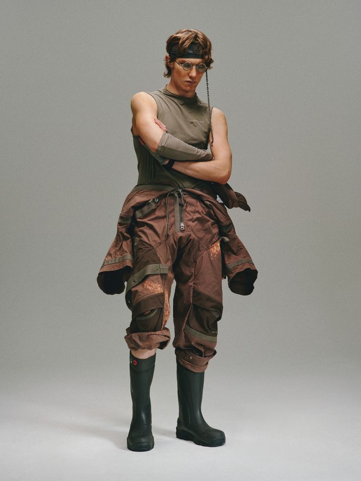 a fullbody studio shot of a male model showcasing a postapocalyptic or utilitarianinspired outfit. the model is wearing a dark brown layered pants, an olive green sleeveless top, black hunterstyle rain boots, and a dark headband. the overall look is highfashion but with streetwear influences.  the background is a muted gray, focusing attention on the model and clothing.  the model’s arms are crossed, presenting a somewhat serious or stoic demeanor. the clothing features design elements suggesting functionality and layering.  the image style is contemporary and stylish.