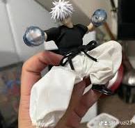 a closeup shot shows a hand holding a small figurine of gojo satoru from the anime jujutsu kaisen. the figurine is depicted in action, wearing a black shirt and white pants and has large translucent blue boxing gloves.  the figurine's hair is styled in a characteristically spiky white manner. the background is blurred but shows a part of a room. the focus is primarily on the figurine and the hand holding it.