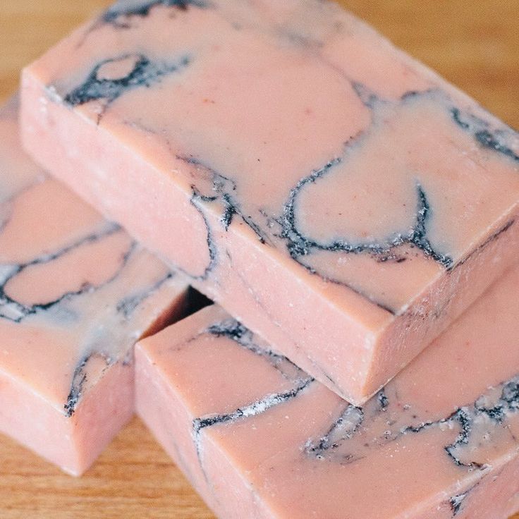 a closeup of three pink soap bars, each with black swirls throughout, the top one is slightly out of focus. they are stacked on top of each other on a wooden surface. the soap looks like it has a natural texture and is very soft and smooth. the black swirls create a unique and luxurious look for the soap.