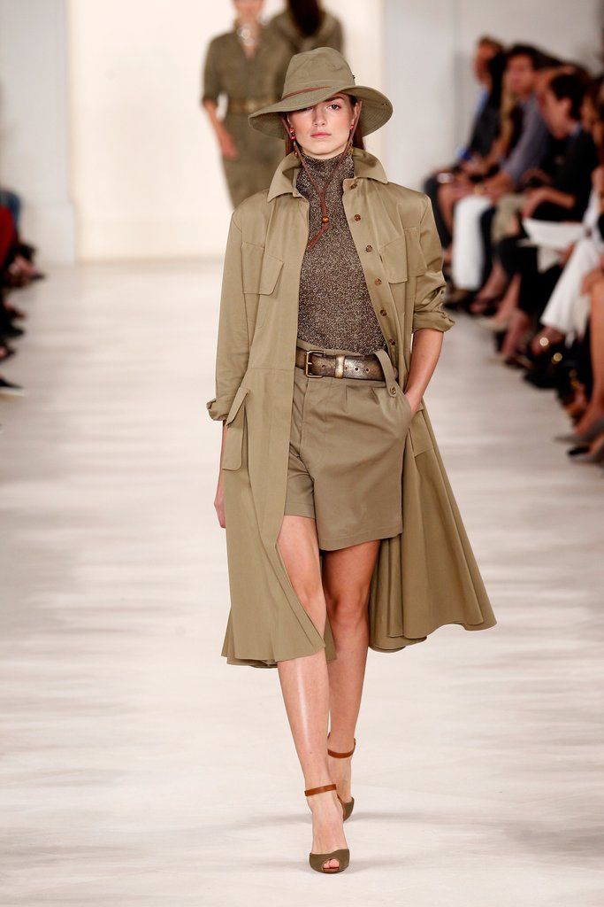 a fashion model walks down the runway wearing a stylish khakicolored outfit. the outfit consists of a long trench coat, khaki shorts, a fitted turtleneck, a brown leather belt, and brown anklestrap sandals. she also wears a khakicolored fedora hat. the background shows other models and spectators. the overall tone is one of high fashion and sophistication.