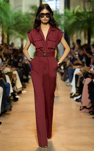 a fashion model walks the runway in a burgundy jumpsuit. she's wearing a belt, sunglasses, jewelry and heels. she has long dark hair. the runway is surrounded by audience members.