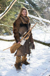 a man dressed in medievalstyle clothing and carrying a bow and arrow stands in a snowy forest. he is wearing a furtrimmed coat, leather boots and pants, and a scarf around his neck. the man has long blond hair and a beard.  the scene is peaceful and evocative of a historical or fantasy setting.