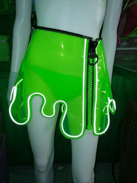 a woman is wearing a neon green skirt with a zipper down the center and a white neon trim. the skirt is transparent, showing her white legs. she is standing in front of a green background. she is wearing a clubwear outfit that is futuristic and sexy.
