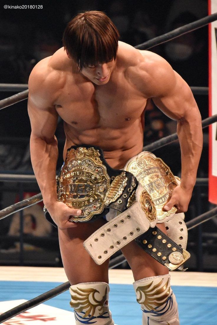 a closeup shot of a muscular male professional wrestler holding two championship belts. he is shirtless and his body is welldefined. he's looking down at the belts, appearing proud and triumphant. the wrestler is wearing wrestling boots with gold accents. the background shows a wrestling ring and some spectators in the background. the photo is welllit and the wrestler's expression conveys satisfaction and accomplishment.