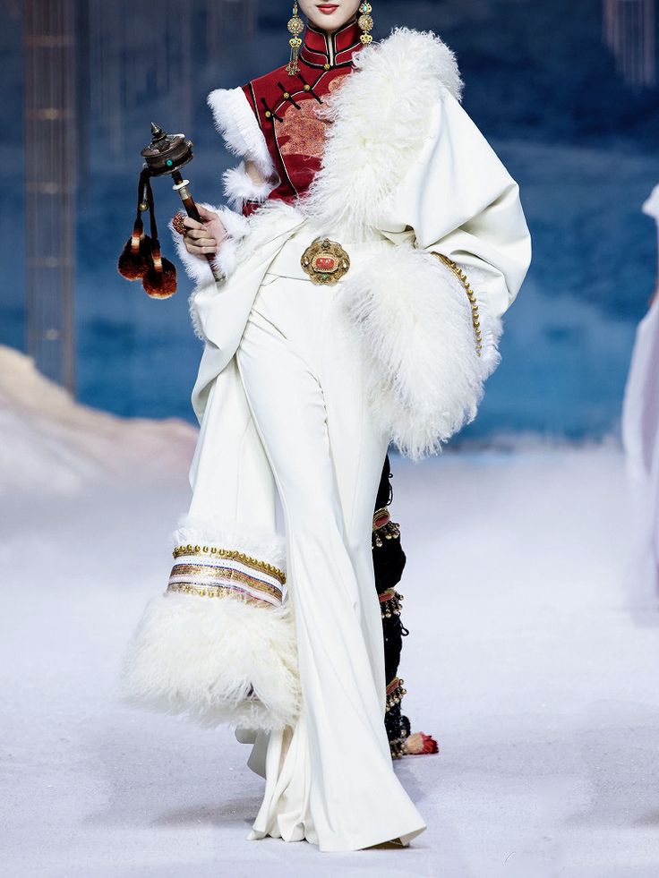 a fashion model walks the runway wearing a white fur coat and matching pants. the coat has gold trim and a red and gold top underneath. the model is wearing a silver belt with a large red stone. she is also wearing a pair of black boots with gold trim. the model is posing for the camera with a confident expression on her face. the runway is white and snowy, suggesting a wintery theme.