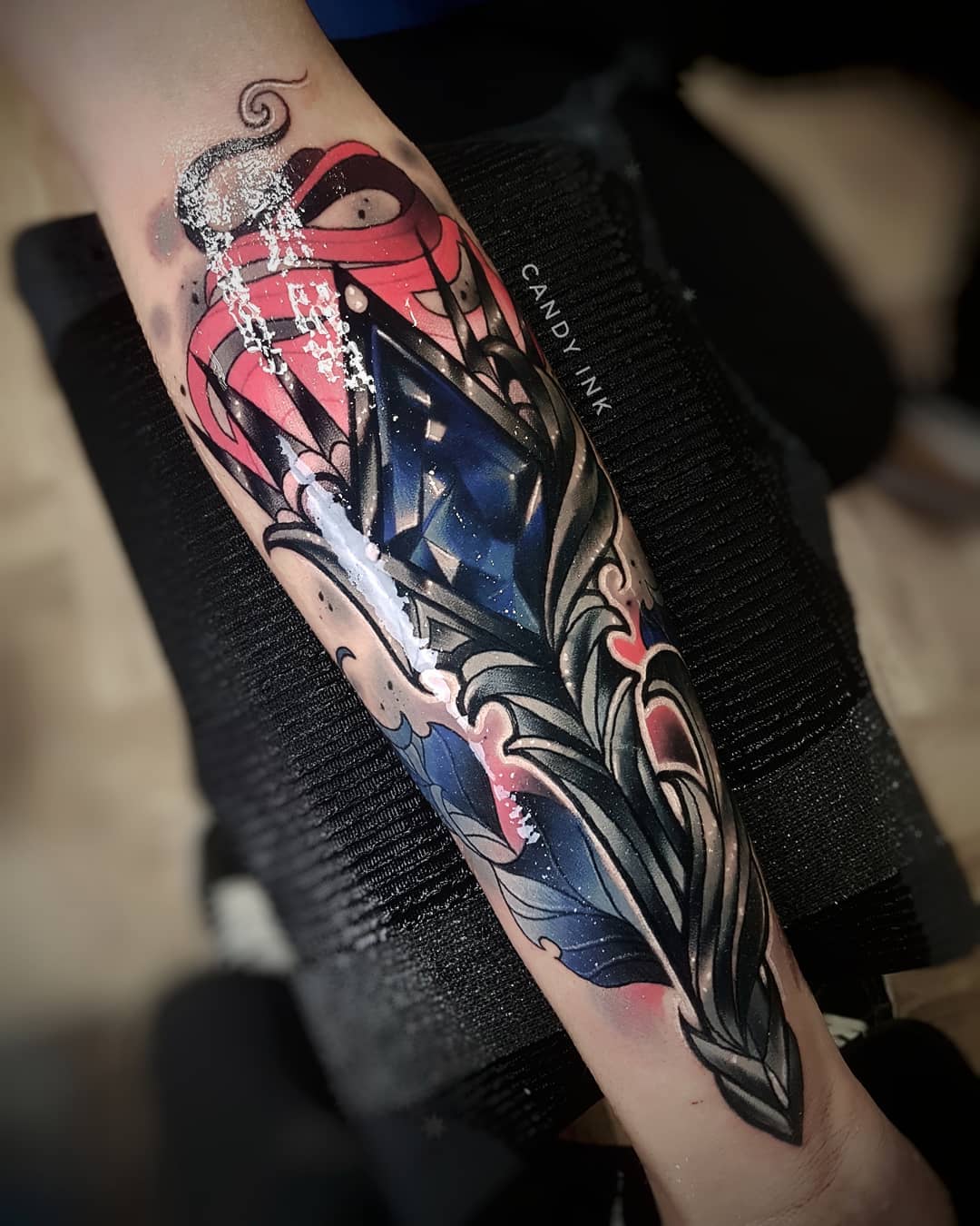 a freshly done tattoo of a blue crystal surrounded by dark gray leaves and red accents. the tattoo is on a person's inner forearm. the tattoo artist used white ink for highlights. the tattoo studio name "candy ink" is visible on the inner bicep.