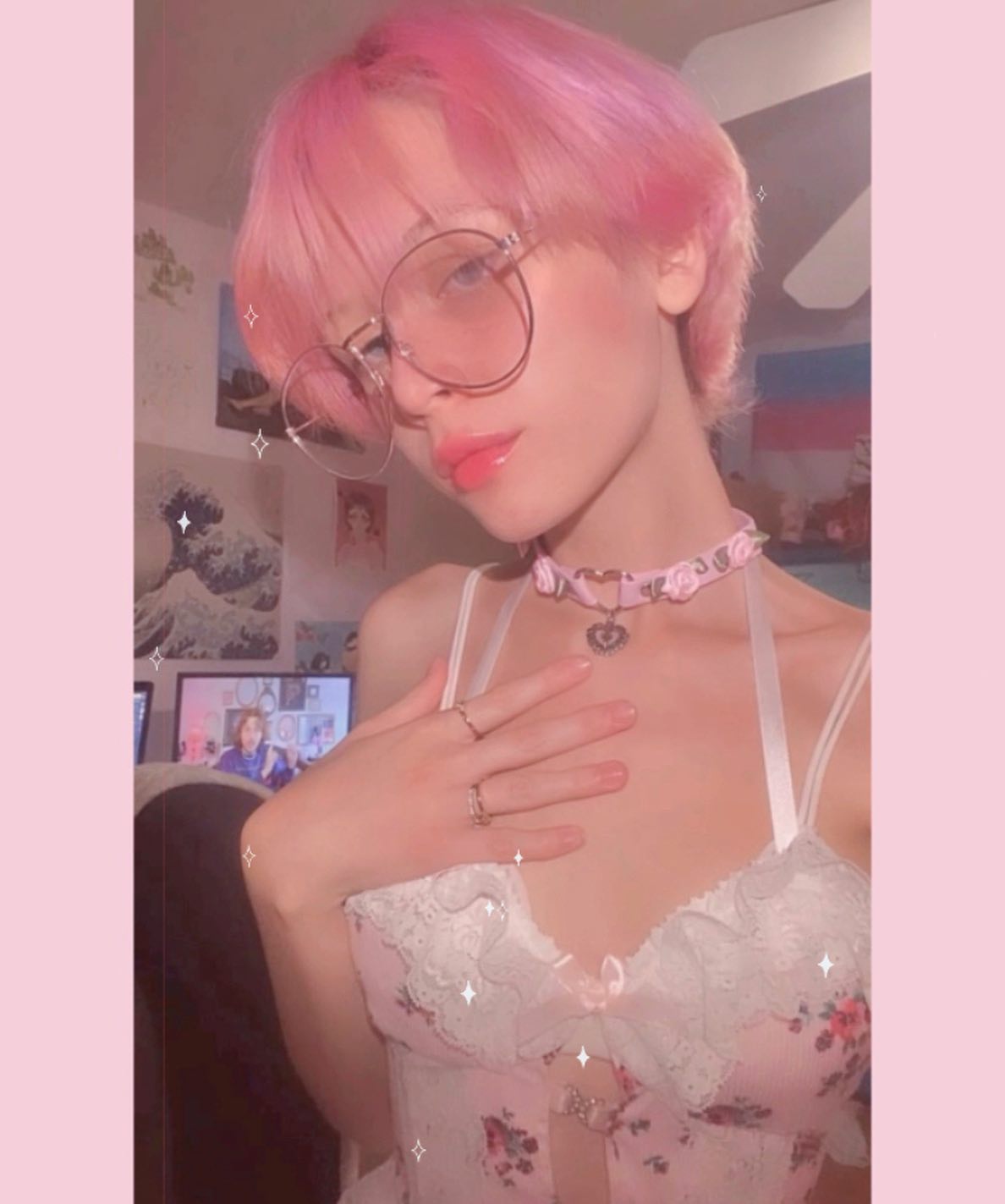 closeup selfie of a young woman with short, bright pink hair. she's wearing round pinkrimmed glasses, a pink floral lace camisole, and a pink choker necklace with a small heart pendant. she has two rings on her left hand. the background is softly lit and features a glimpse of a computer screen, an animestyle poster, and other bedroom details. the overall aesthetic is very pastel and cute, reminiscent of kawaii style.  the image has a soft, filtered look.