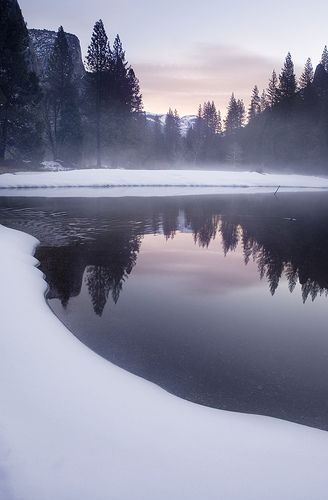 a serene winter landscape features a calm lake reflecting the surrounding forest of tall trees and snowcovered banks. mountains loom in the background beneath a soft, overcast sky. the scene is tranquil, with a cold, peaceful atmosphere.
