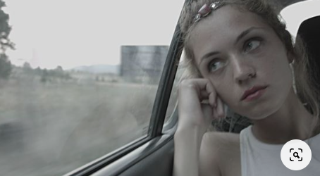 a young woman with blonde hair and a nose ring sits pensively in the back seat of a car, gazing out the window at a passing landscape. the image is in grayscale, giving it a melancholic and introspective feel. the woman's expression suggests she is lost in thought, perhaps reflecting on her journey or the destination ahead.