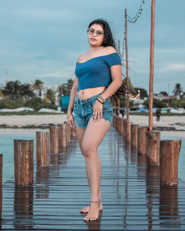 a young woman with long dark hair is standing confidently on a wooden pier overlooking a calm body of water. she's wearing a blue offtheshoulder top, denim shorts, and sunglasses. her casual attire and relaxed pose suggest a summer vacation or beach getaway. the setting appears tranquil and serene, with a clear sky and a peaceful ocean in the background. the overall impression is one of carefree summer style and natural beauty.