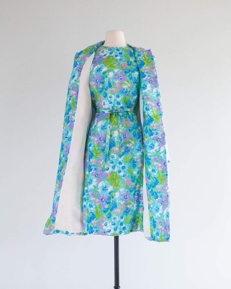 a vintage floral dress with a matching coat is displayed on a mannequin. the dress features a blue, purple, and green floral pattern, has a shift silhouette and is belted at the waist. a matching coat with a white lining is draped over the dress. the ensemble appears to be from the 1960s era.
