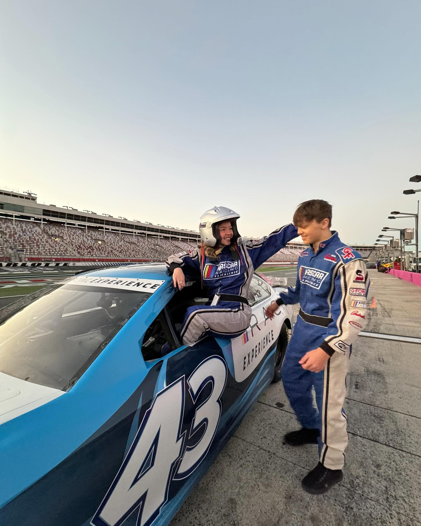 a young woman and man in matching nascar racing suits stand next to a blue #43 race car. the woman is sitting in the car and the man is standing next to her. they are both smiling. the background shows a race track.