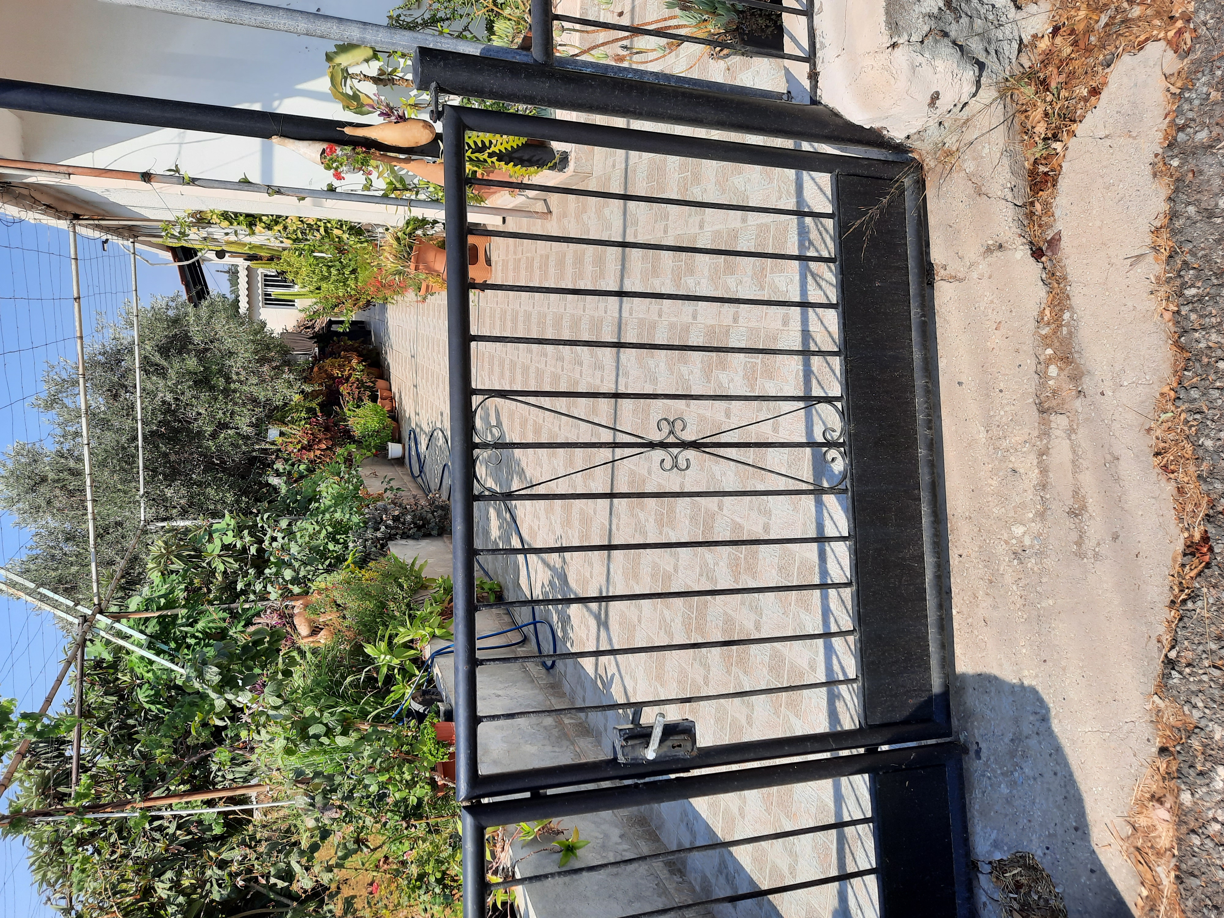 a black wrought iron gate leads into a garden filled with lush green plants and a paved pathway. the garden appears wellmaintained and leads towards a house.