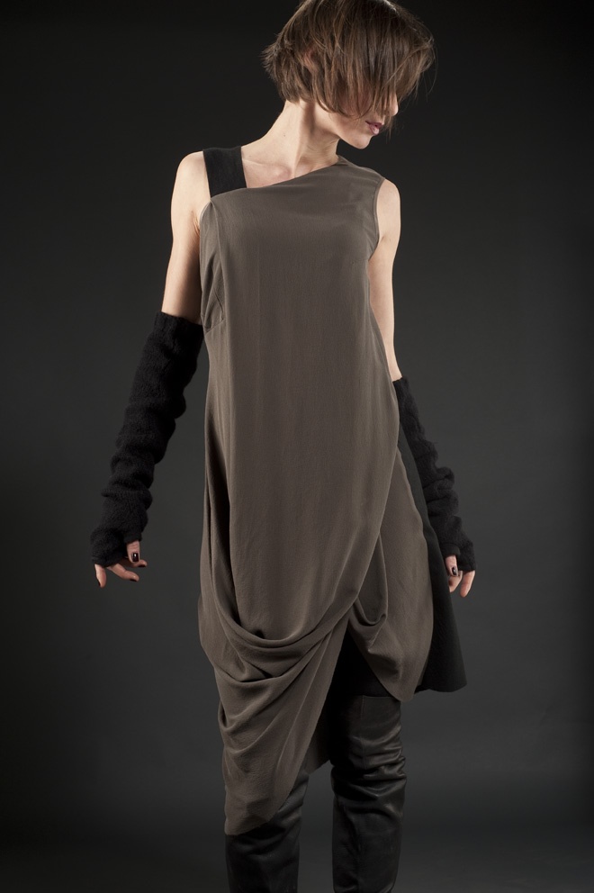 a young woman in a long grey dress with black accents poses against a dark background. she is wearing black long sleeves, has a casual hairstyle, and is looking down towards the right. the image has a fashion photography style and a minimalist feel.