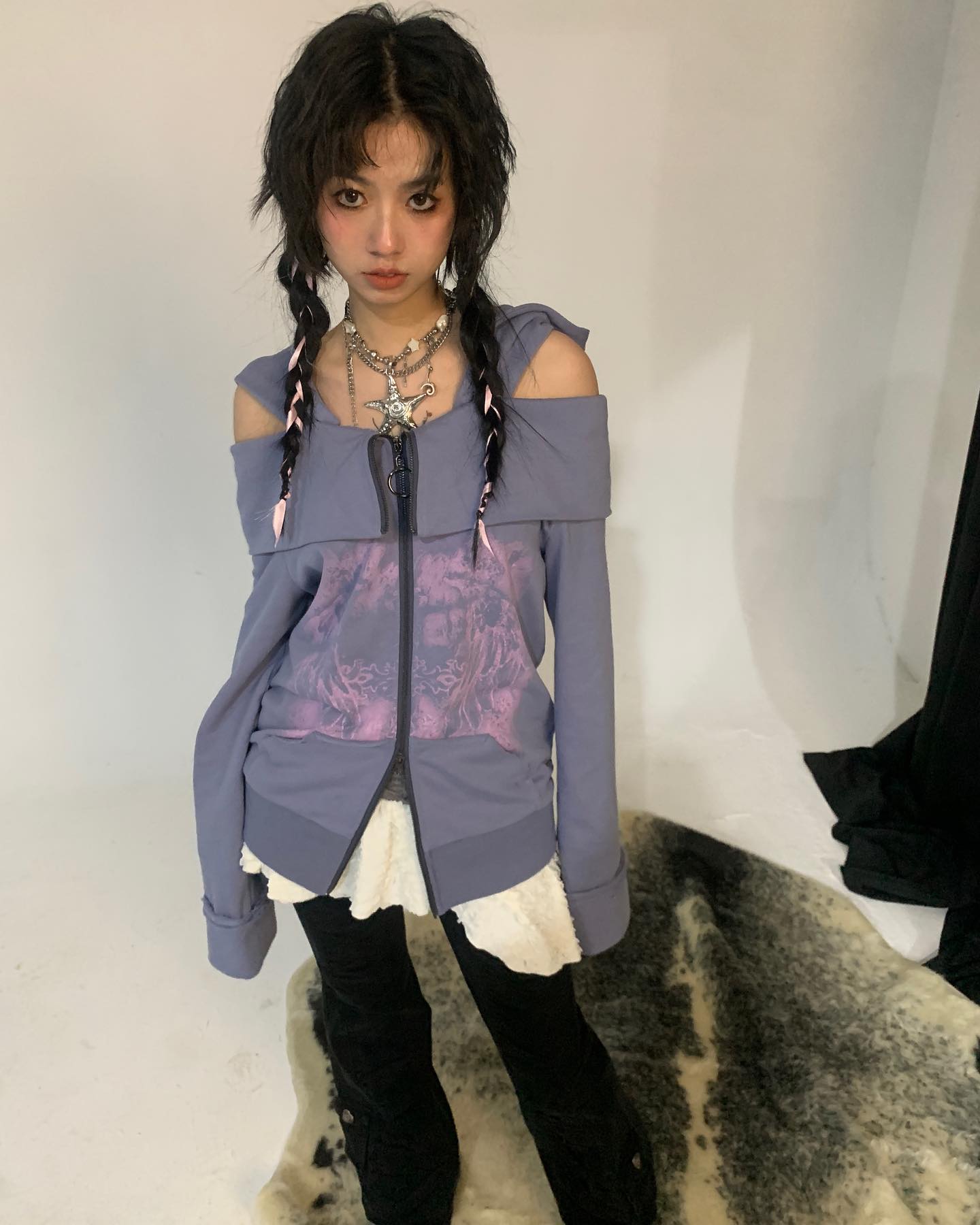 a young woman with dark hair styled in pigtails poses in a studio wearing a purple offshoulder zipup sweater with a graphic print, black corduroy pants, and silver necklaces.  she is standing on a fluffy rug. the overall style is grunge and alternative.