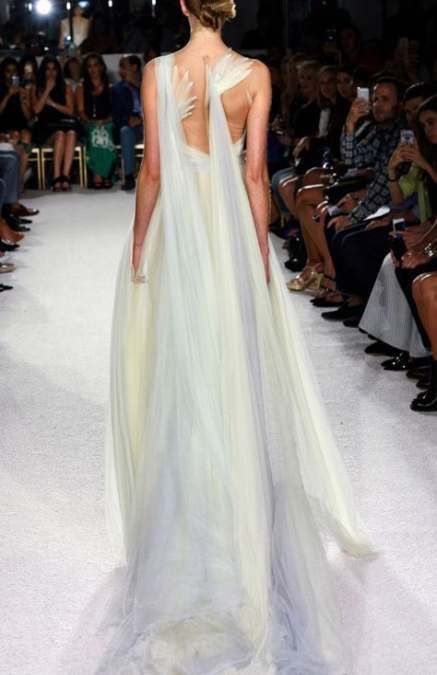 a woman in a white gown with a long flowing tulle skirt walks the runway. she is posing for the camera with her hair pulled back and a simple white dress. the dress is sheer and you can see her back and shoulders. the runway is white and there are people in the background, blurred.