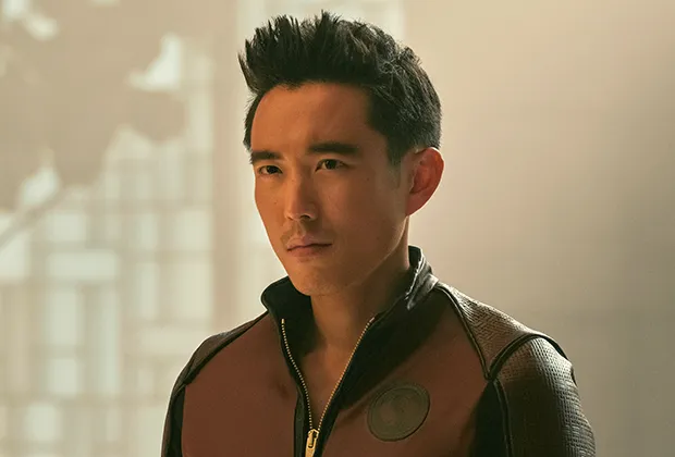 a headshot of a man with short black hair and a maroon leather jacket. he has a serious expression on his face. the background is blurry and appears to be an interior setting.