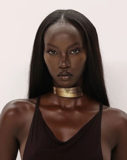the image is a closeup portrait of a young black woman with long, straight black hair. she is wearing a simple, darkcolored sleeveless top and a striking gold multilayered choker necklace. her expression is serious and her makeup is minimal, highlighting her flawless dark skin. the background is plain white, keeping the focus entirely on the model. the overall aesthetic is clean, elegant, and sophisticated.