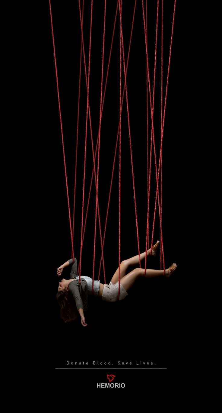 a woman is hanging from red strings against a black background. the woman is tied up by her hands and feet. the image is meant to be dramatic and raise awareness about the importance of donating blood. the text "donate blood. save lives." is written below the image. the image is intended to be eyecatching and make people think about the importance of blood donation.