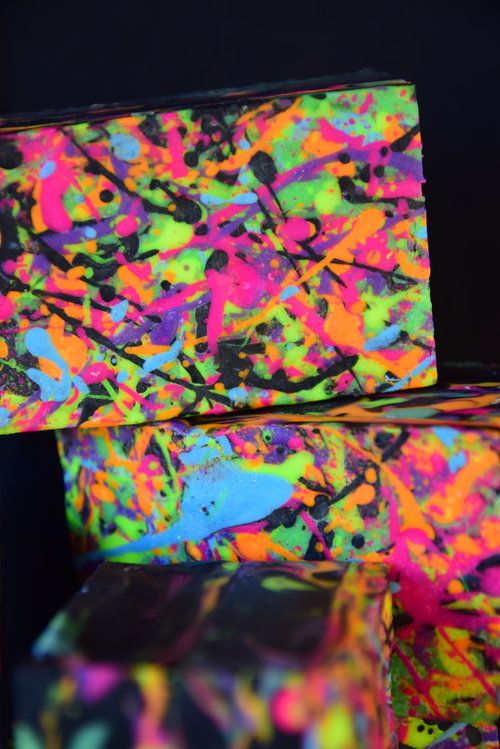 a closeup shot of a stack of three brightly colored soap bars with a neon paint splatter design. the soaps are stacked on top of each other, and the paint splatter design is visible on all three. the colors are bright and vibrant, and the design is messy and chaotic. the background is a dark, black, and the colors of the soaps pop out from the background. the image is dynamic and energetic.