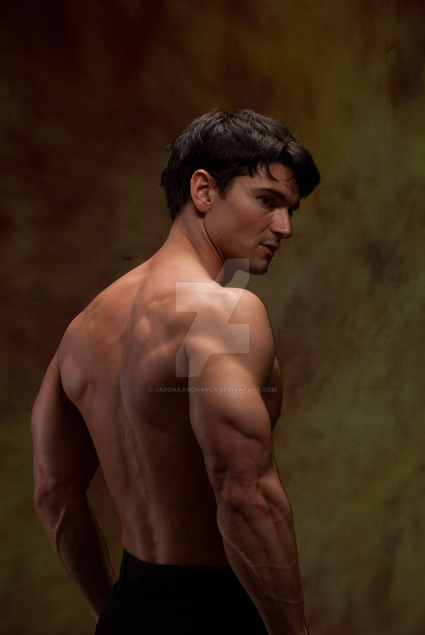 a muscular man stands shirtless, looking away from the camera, with his back turned to the viewer. his back, shoulders, and arms are visible, showcasing his physique. the man is in a studio setting.