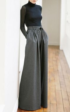 a woman is standing in a room with a white wall, wearing a black turtleneck and gray, wideleg pants with a black belt. her outfit is stylish and minimal. the woman is looking at the camera.