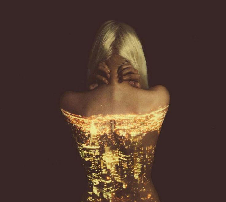 the image showcases a blonde woman standing with her back to the camera. her hands are gently placed on her neck.  a projection of a glittering cityscape in warm golden tones is mapped onto her back, making it appear as if the city is integrated into her body. the background is dark and indistinct, drawing all attention to the striking contrast between the woman's skin and the projected light of the city. the overall effect is surreal and artistic, combining elements of human form and urban landscape in a visually arresting manner.