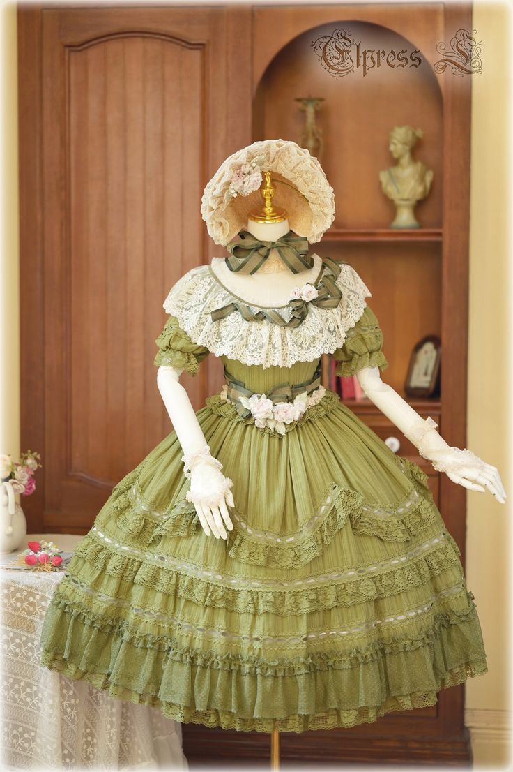 a green lolita fashion dress is displayed on a mannequin in front of a wooden door. the dress features a square neckline with lace trim, puffed sleeves, and a tiered skirt with ruffles and lace detailing. a matching bonnet with flowers rests on the mannequin's head. the background features a shelf holding a bust and a picture frame. a tea set is partially visible on the left. the overall aesthetic is romantic and vintageinspired.