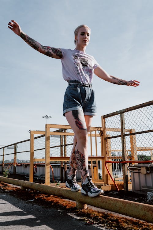 a young woman with tattoos on her arms and legs balances on a metal rail in an urban setting.  she's wearing denim shorts, a light purple tshirt, and sneakers. the background features a chainlink fence and industrial structures under a bright, sunny sky. the overall mood is casual and athletic.