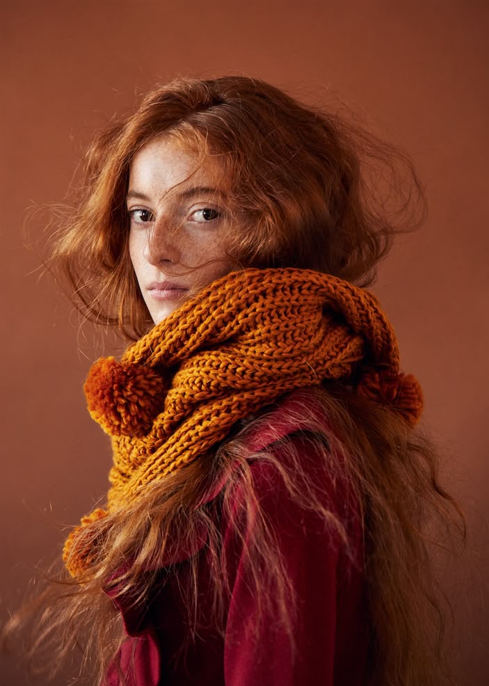 a portrait of a young woman with red hair and freckles, looking at the camera. she is wearing a chunky knitted scarf in autumn colors with pompoms, and a red shirt. the background is a solid brown. the image is a studio shot.