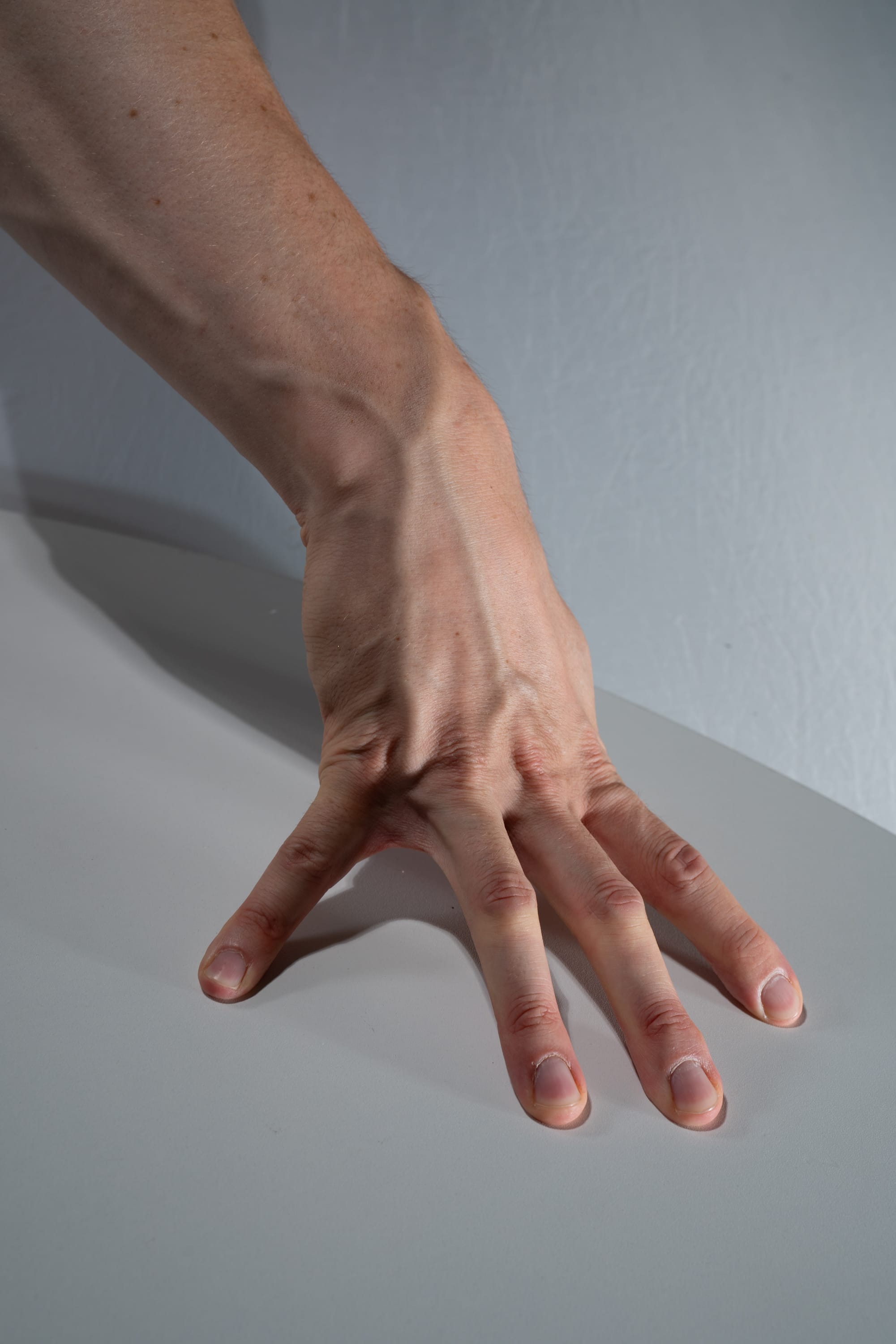 a closeup side view of a man's hand and arm resting on a neutral gray surface. the image highlights the details of the skin, veins, and muscles of the hand and arm. the studio lighting emphasizes the contours and textures of the subject.