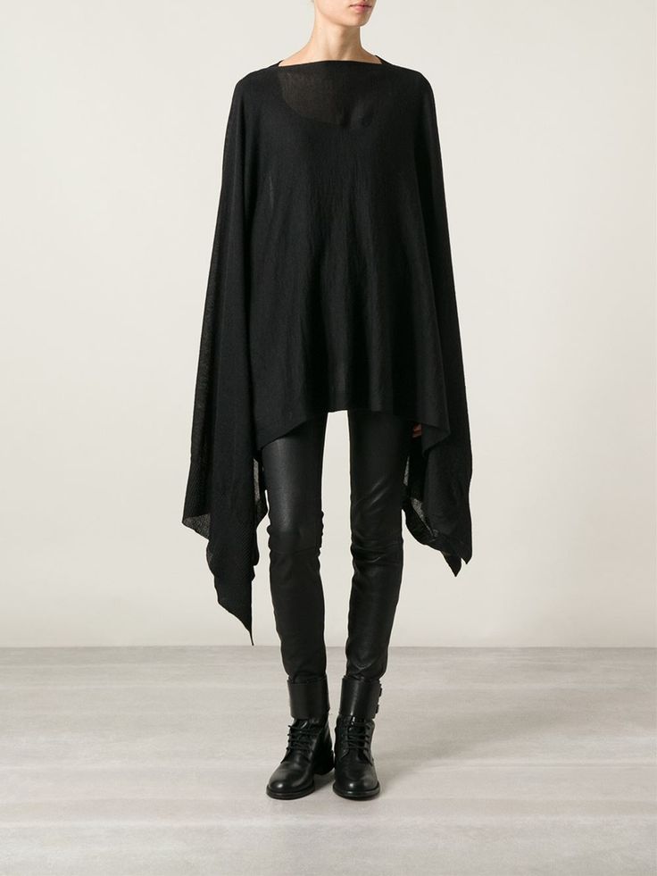 a woman stands in front of a white wall wearing a black poncho, black leggings, and black boots. she is centered in the frame, facing forward. the poncho is oversized with long, draped sleeves and an uneven hemline. the leggings are tightfitting, and the boots are laceup style. the overall look is modern and minimalist. the background is plain and neutral, which helps to emphasize the woman and her outfit.