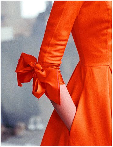 a closeup shot of the sleeve of an orange dress, adorned with a large bow at the cuff. the dress has a structured bodice and flowing skirt. the arm is visible in the sleeve, but most of the body is out of frame.