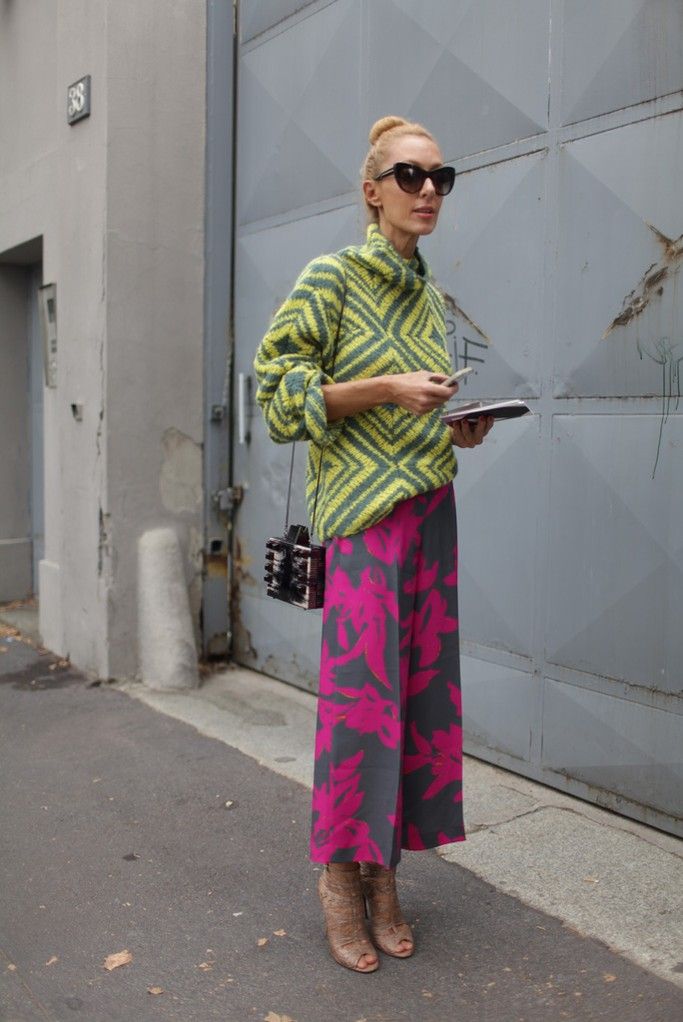 a stylish blonde woman is standing in front of a grey metal gate. she is wearing a vibrant green and yellow geometric patterned sweater, fuchsia and grey floral printed wideleg pants, and brown ankle boots with heels. she carries a small black rectangular bag and is wearing sunglasses. the overall look is chic and trendy, typical of street style fashion.